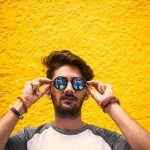 Young man with sunglasses standing against a vibrant yellow wall in daylight.